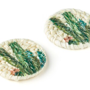 Puede incluir: Dos botones redondos tejidos de color blanco con un diseño floral verde y rosa. Los botones están hechos de materiales naturales y tienen un aspecto rústico y hecho a mano.