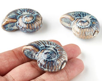 May include: Three ceramic ammonite fossils with a spiral design. The fossils have a colour palette of brown, blue, and cream. One fossil is held in a hand. The fossils are decorative and can be used as home decor.