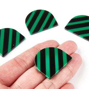 Könnte beinhalten: Vier schwarz-grün gestreifte, halbkreisförmige Anhänger mit zwei kleinen Löchern. Die Anhänger werden in einer Hand vor weißem Hintergrund gehalten. Die Anhänger haben eine glänzende Oberfläche und eine abgerundete Oberseite.