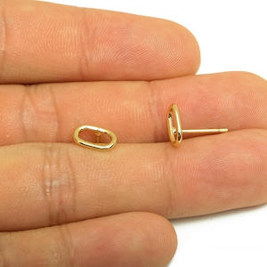 以下が含まれることがあります： 2つの金色の楕円形のスタッドピアス。片方のピアスはシングルスタッド、もう片方はペアです。ピアスは小さく、シンプルでミニマリストなデザインです。ピアスは人の手で表示されています。