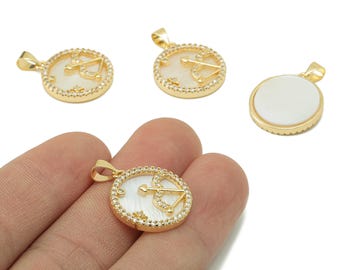 May include: Gold-colored pendant charms with a white center and a bow and arrow design. The charms are round with a decorative border of small, clear stones. One charm is held in a hand, while others are scattered on a white surface.
