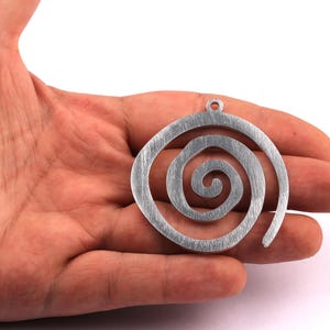 Puede incluir: Colgante plateado en forma de espiral con superficie texturizada. El colgante tiene un pequeño lazo en la parte superior para colgarlo. El diseño presenta una serie de curvas concéntricas, creando un patrón arremolinado. Esta joya es adecuada para collares.