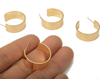 May include: Gold-colored hoop earrings with a flat, wide design. The earrings are a simple, modern style, with a post for insertion into the ear. Several earrings are visible, with one held in a hand.