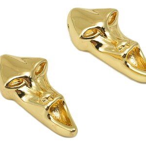 May include: Two gold-colored metal face mask charms with a detailed design. The masks have a wide open mouth and a stylized face.