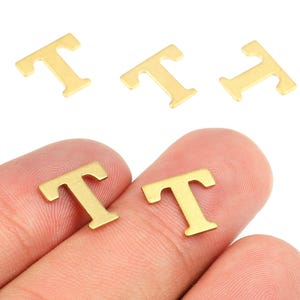 Puede incluir: Amuletos de metal dorado con forma de letra "T". Se ven cinco amuletos, dos de ellos sostenidos entre los dedos. Los amuletos son pequeños, planos y tienen un diseño sencillo sin serifa. Adecuados para la fabricación de joyas o proyectos de artesanía.