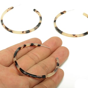 May include: A pair of hoop earrings with a tortoiseshell pattern. The earrings are a light beige color with dark brown and black spots. One earring is held in a hand, and two others are displayed on a white background.