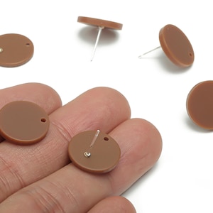 Könnte beinhalten: Braune, kreisförmige Ohrstecker mit silbernen Stiften. Sie werden auf weißem Hintergrund präsentiert, einige werden in einer Hand gehalten. Jeder Ohrring hat ein kleines Loch für den Stift.
