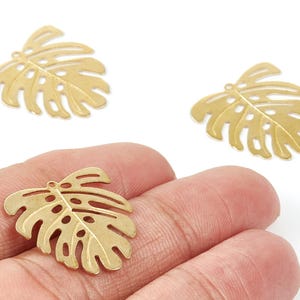 Puede incluir: Tres dijes dorados con forma de hoja de Monstera. Los dijes tienen un diseño calado detallado con múltiples agujeros y una vena central. Un dije se sostiene en una mano, mientras que otros dos se muestran sobre un fondo blanco. Estos dijes son adecuados para la fabricación de joyas.