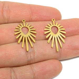 May include: Two gold-coloured sunburst-shaped pendants. Each pendant has a central teardrop shape with radiating lines, and a small loop at the top for attaching to a chain or earring hook. The pendants are held in a hand.