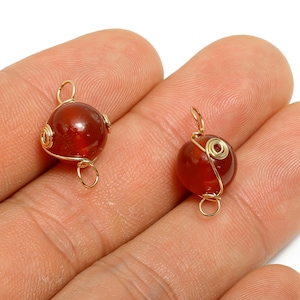 May include: Two round, reddish-brown gemstone pendants with gold-colored wire wrapping. The wire is coiled around the stones and forms a loop at the top and bottom for attaching to a chain or earring hook. The background is a light color.