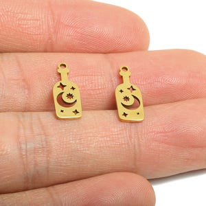 May include: Two small, gold-coloured bottle charms with a celestial design. Each charm features a bottle shape with a crescent moon, stars, and a small hole at the top for stringing. The charms are held between fingers.