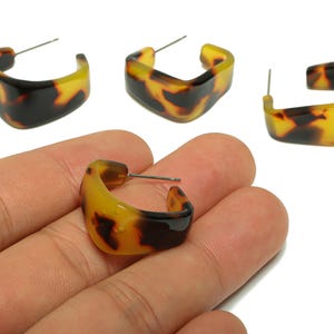 May include: Four square-shaped hoop earrings with a tortoiseshell pattern. One earring is held in a hand. The earrings have a post for pierced ears. The earrings are yellow, brown, and black.