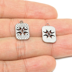 Puede incluir: Dos colgantes rectangulares plateados con diseños decorativos. Uno presenta un patrón detallado de líneas y formas, el otro un diseño más simple y recortado. Ambos tienen pequeños bucles para sujetar.