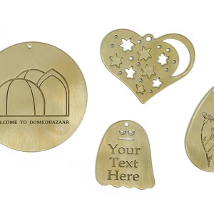 Puede incluir: Cuatro adornos de latón con diferentes diseños. Uno es un círculo con un edificio en forma de cúpula y el texto "WELCOME TO DOMEDBAZAAR". Otro es un corazón con estrellas y una luna creciente. El tercero es una forma de fantasma con el texto "Your Text Here". El último es una forma de lágrima con un diseño floral.