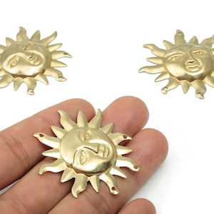 Könnte beinhalten: Goldfarbene sonnenförmige Charms mit einem lächelnden Gesicht und strahlenden Strahlen. Die Charms haben kleine Löcher zum Befestigen an Schmuck oder anderen Bastelarbeiten. Drei Charms sind sichtbar, einer wird in einer Hand gehalten.