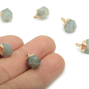 May include: A collection of small, pale green gemstone pendants with gold-coloured metal detailing. The pendants are hexagonal and have a loop for attaching to jewellery. The gemstones are held in a hand.