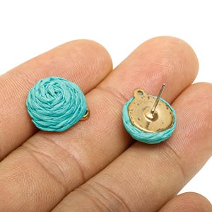 May include: A pair of turquoise blue stud earrings. Each earring has a woven, circular design with a gold-coloured post and backing. The earrings are held in a person's hand against a white background.