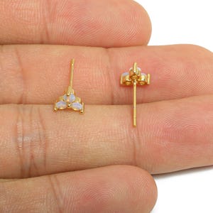 May include: Two gold-colored stud earrings, each featuring a cluster of three oval, opalescent stones. The earrings are displayed against a skin-toned background, showcasing their delicate design and color.