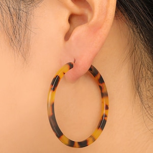 May include: A close-up of a tortoise shell hoop earring. The earring is a large, circular design with a warm, brown and yellow pattern. The earring is worn on the ear, with the post inserted through the earlobe.