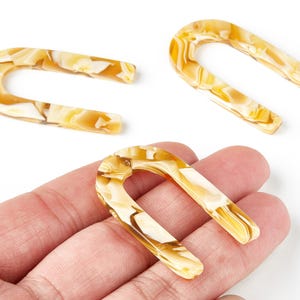 Può includere: Tre fermagli per capelli a forma di U con un motivo marmorizzato nei colori giallo e crema. Un fermaglio è tenuto in mano, mentre gli altri due sono su sfondo bianco. I fermagli sono realizzati in materiale lucido.