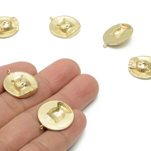 Puede incluir: Amuletos dorados con forma de gato. Los amuletos son redondos con un diseño de cara de gato en relieve. Cada amuleto tiene un pequeño lazo en la parte superior para sujetarlo a la joyería. Los amuletos se muestran sobre un fondo blanco.