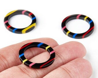 May include: Three black resin rings with colorful stripes of red, yellow, pink, and blue. The rings are circular with a flat surface and are shown against a white background. One ring is held in a hand.