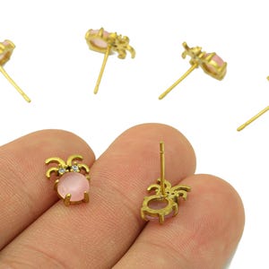 Puede incluir: Pendientes de botón dorados con una piedra rosa ovalada y pequeños cristales transparentes. Los pendientes tienen un diseño decorativo alrededor de la piedra rosa. Se ven varios pendientes, algunos enfocados y otros borrosos.