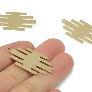 May include: Three gold-coloured metal pendant charms. Each charm has a rectangular shape with horizontal bars extending from the sides. One charm is held in a hand. The charms have small holes for attaching to jewellery.