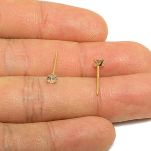 Puede incluir: Dos pendientes de botón dorados con piedras preciosas redondas y transparentes. Los pendientes se sostienen en una mano sobre un fondo blanco. Las piedras están engastadas en un engaste de cuatro puntas.