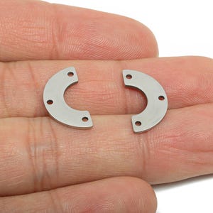 May include: Two silver-coloured, semi-circular metal components with three small holes each. The pieces are held in a hand, and are likely jewellery findings or craft supplies.