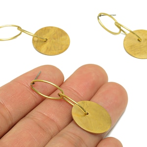May include: Gold-colored dangle earrings. Each earring features a small oval loop connected to a circular disc. The earrings have a post for pierced ears. The earrings are made of a shiny, metallic material.