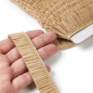 Puede incluir: Primer plano de un rollo de flecos beige con borde dorado. Una mano sostiene un trozo de los flecos, mostrando su textura. Los flecos están hechos de un material tejido, perfecto para proyectos de manualidades.