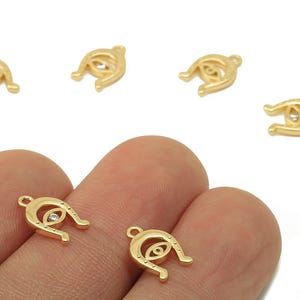 May include: Gold-colored charm pendants in the shape of an eye, with a horseshoe-like design. Each charm features a small, clear crystal in the center of the eye. The charms are small, with a loop at the top for attaching to jewelry.