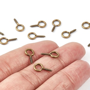 Peut inclure: Une collection de petits œillets à vis de couleur laiton antique. Ces œillets ont une base filetée et une boucle circulaire pour la fixation à divers projets d'artisanat. Les œillets sont dispersés sur une surface blanche.
