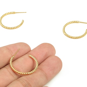 May include: Gold-coloured hoop earrings with a twisted rope design. The earrings are shown on a white background, with one held in a hand. The earrings have a post closure. The earrings are a jewelry item.