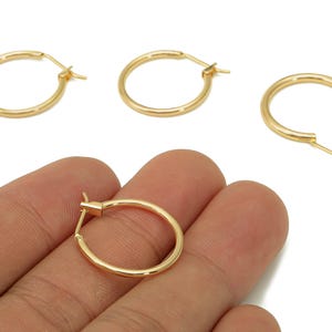 May include: Gold-colored hoop earrings. The earrings are round and have a simple, classic design. One earring is being held by a hand. The earrings are made of a shiny, metallic material. The image is taken against a white background.