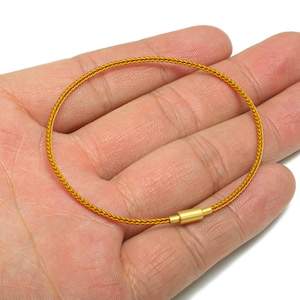 Puede incluir: Una pulsera dorada con un diseño tejido. La pulsera es circular y tiene un pequeño cierre rectangular. La pulsera se sostiene en una mano y el fondo es blanco.