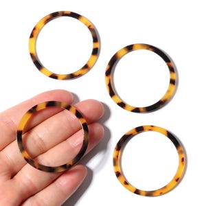Op de afbeelding: Vier cirkelvormige harsringen met een schildpadpatroon in amber en donkerbruin. Eén ring wordt in een hand gehouden, de andere liggen op een wit oppervlak. Deze ringen zijn geschikt voor het maken van sieraden.