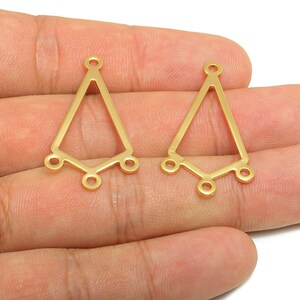 以下が含まれることがあります： 2つの金色の金属製イヤリングコネクタ。各コネクタは、上部にループ、下部に3つのループが付いた三角形の形状をしています。コネクタは肌色の背景に対して表示されています。