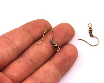 May include: Two antique bronze-colored earring hooks. One hook is in focus, showing a curved wire, a coiled spring, and a small bead. The other hook is slightly out of focus. The hooks are on a white background.