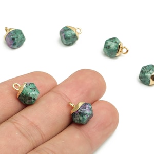 Può includere: Ciondoli in pietra preziosa verde e viola con cappucci e anelli color oro. I ciondoli sono di forma irregolare e sembrano essere pietre naturali. Diversi ciondoli sono sparsi su una superficie bianca.