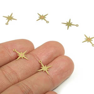 May include: Several small, gold-colored starburst charms with a small pink center and a loop at the end. The charms are scattered on a white background, with some held in a person's hand. These charms are ideal for jewelry making.