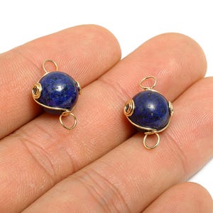 May include: Two round, deep blue lapis lazuli beads with gold-colored wire wrapping. Each bead has a small loop at the top and bottom for stringing. The beads are shown against a neutral background.