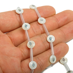 May include: A close-up of a necklace featuring white, circular beads with a light blue design, strung on clear, thin strands. The necklace is held in a person's hand against a white background, showcasing the delicate design.