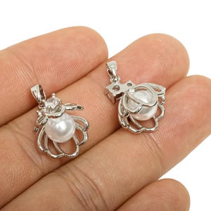 May include: Two silver-colored pendants, each featuring a white pearl nestled within a stylized, openwork design. One pendant has a small, clear gemstone accent. The pendants are held in a hand.