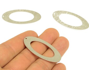 May include: Three silver-colored oval-shaped metal jewelry findings. One finding is held in a hand, showing a small hole at the top. The other two findings are on a white background. The findings have a brushed metal texture.