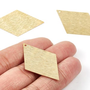 May include: Three gold-colored, diamond-shaped metal pendants with a textured surface. One pendant is held in a hand, while two others are positioned above. Each has a small hole at the top for stringing.