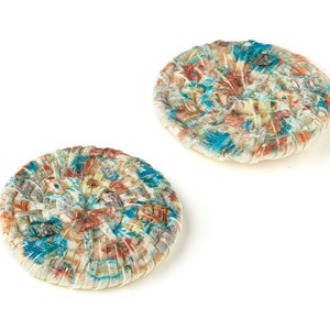 Puede incluir: Dos botones redondos tejidos con una base blanca y un patrón colorido de azul, rojo y amarillo. Los botones están hechos de tela y tienen una superficie texturizada.