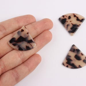 May include: Three fan-shaped, tortoiseshell-patterned beads. One bead rests in a hand, while two others are positioned on a white surface. The beads have a light beige base with dark brown and black mottled patterns.