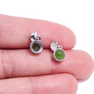 Puede incluir: Un par de pendientes de plata. Un pendiente presenta una piedra preciosa verde, mientras que el otro está vacío. Ambos pendientes tienen una pequeña piedra roja y una parte superior decorativa plateada.